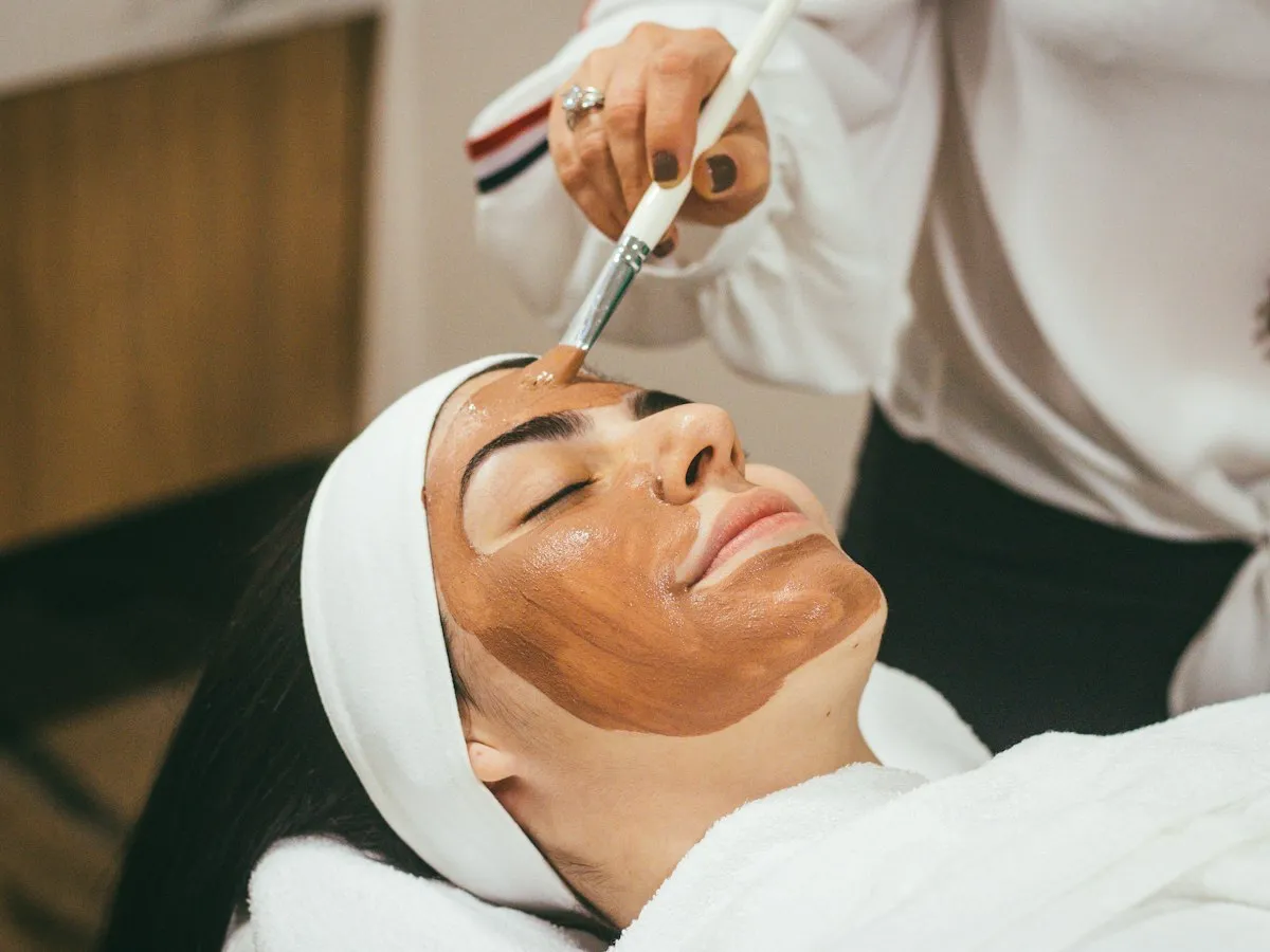 Cosmeticiană care aplică o mască facială cu pensula, clienta stând relaxată cu ochii închiși